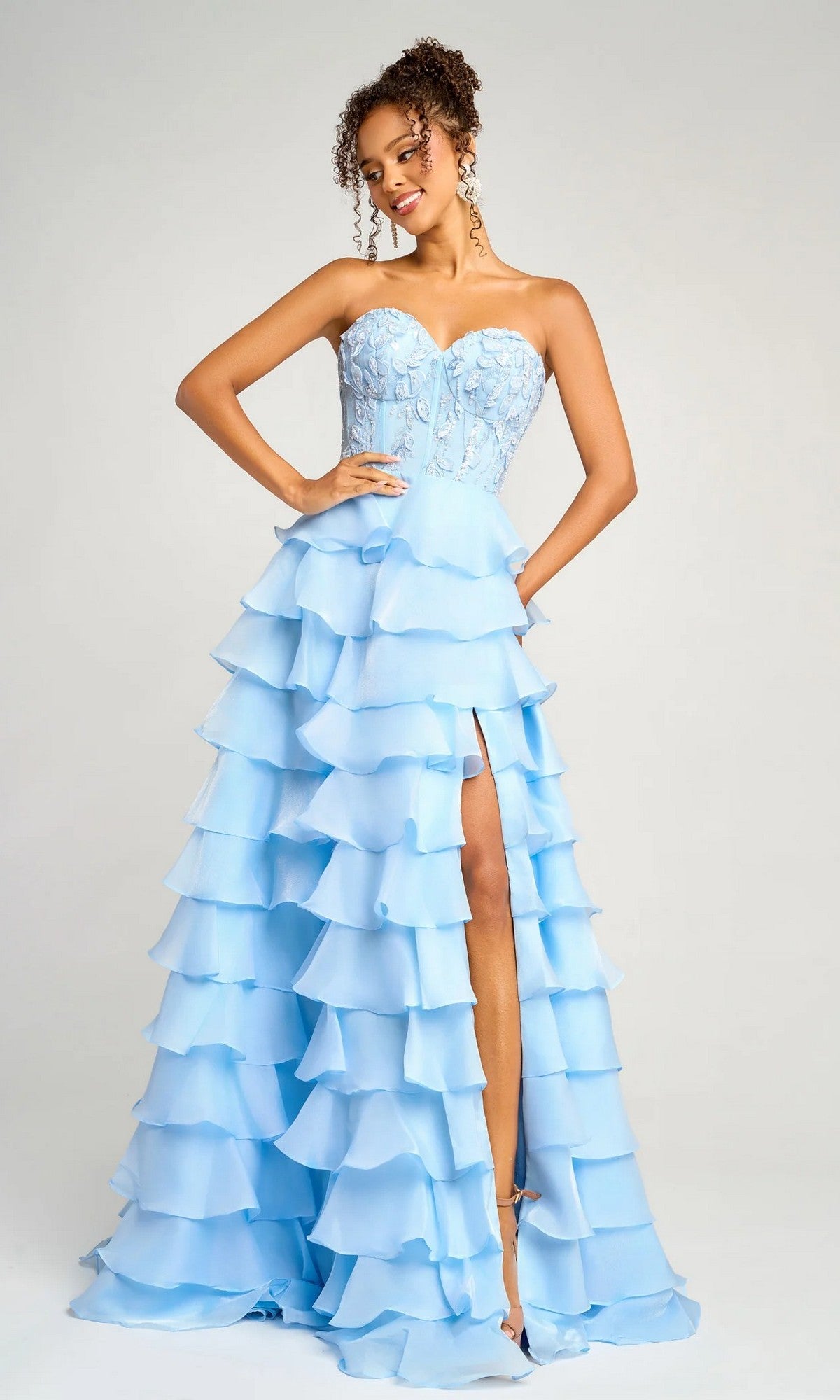 A woman wears the Portia and Scarlett PS26048 long formal dress in blue, featuring a strapless flowy gown with a ruffled tiered skirt, embroidered corset bodice, and thigh-high slit, posing against a plain background.